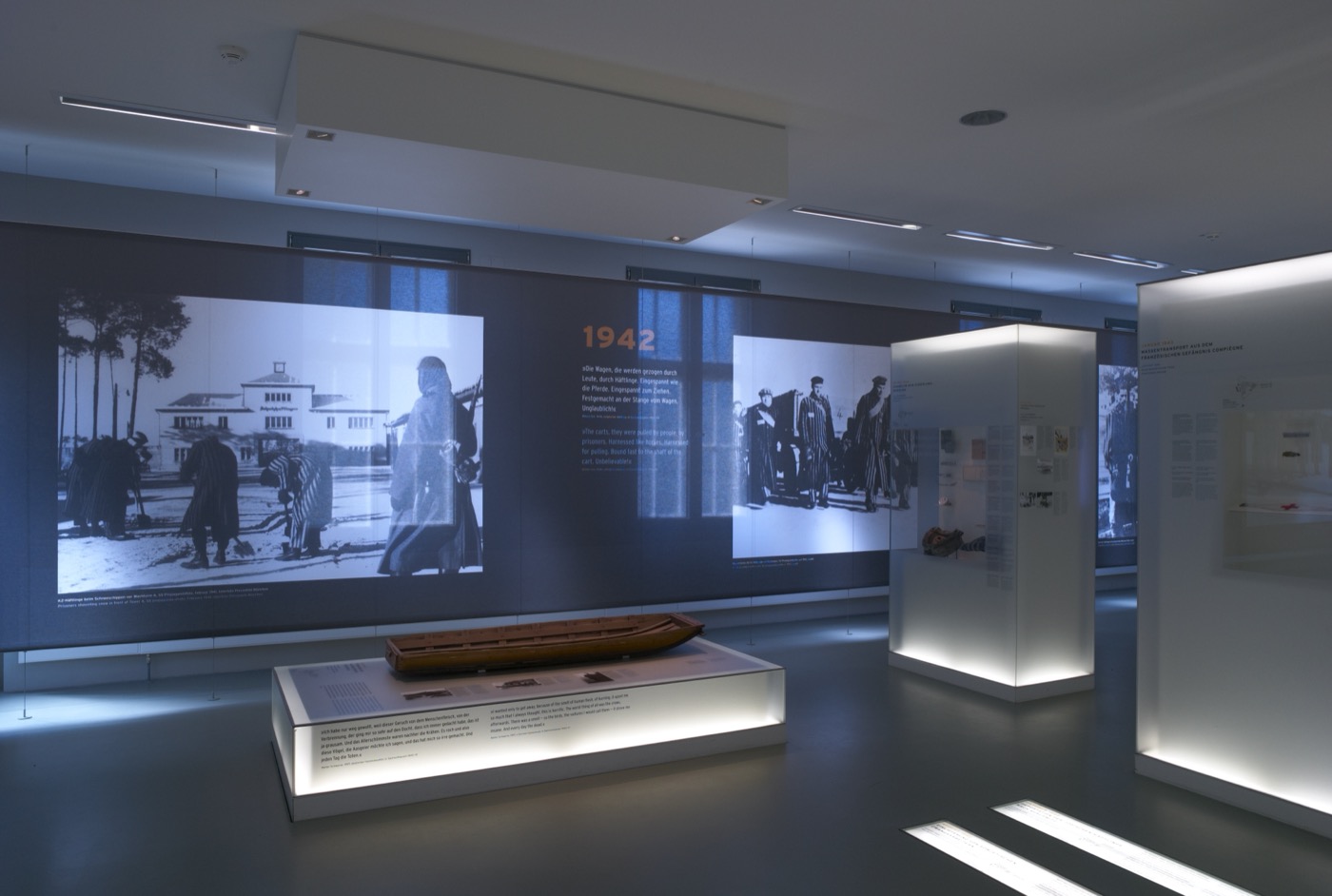 Gedenkstätte Sachsenhausen, Ausstellungsraum mit beleuchteten Vitrinen und bedrucktem Textil an Wandflächen, die beleuchtet sind